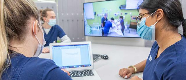 两个戴着口罩的医护人员在笔记本电脑前讨论工作, 在后台有一个屏幕显示正在进行的医疗培训或模拟会议.